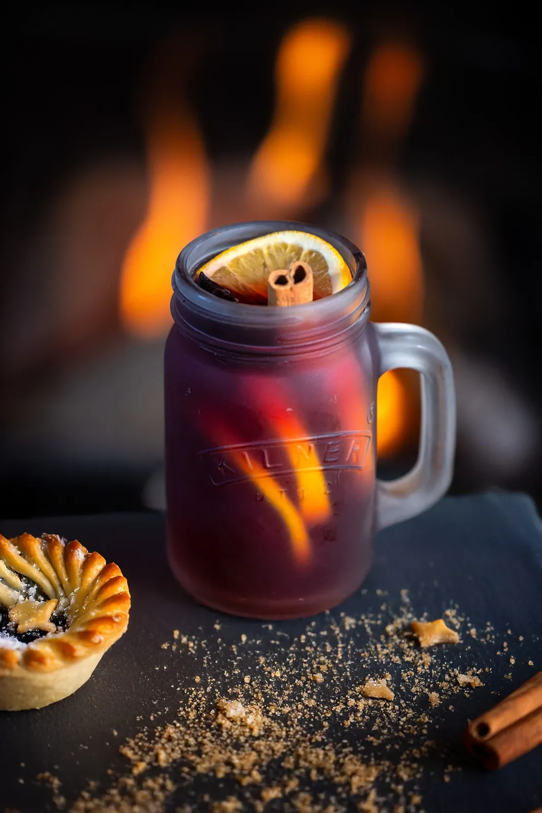 A mulled wine sits in front of a fire with mince pies and cinnamon sticks